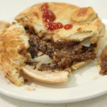 A mince beef and onion pie sitting on a white plate, with the contents of the pie visible. - Thumbnail Image