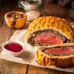 Beef Wellington sitting on some parchment paper, next to small dishes containing sauce and seasoning. - Thumbnail Image
