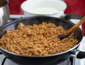 Mince is cooking in a black frying pan, which is sitting on a stove. Next to it is a white saucepan.